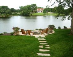 a fire pit in the middle of a grassy area next to a lake with chairs around it