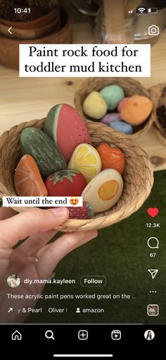 a person holding a basket full of fake food in front of a screen with the words paint rock food for toddlers mud kitchen