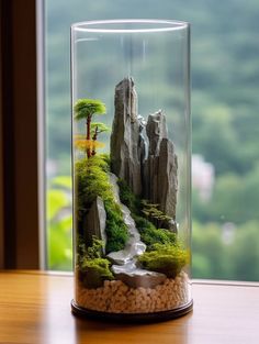 a glass vase filled with rocks and plants on top of a wooden table next to a window