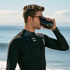 a man in a wet suit drinking from a water bottle while standing on the beach