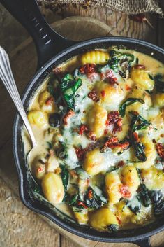 a skillet filled with pasta and spinach on top of a wooden table next to utensils