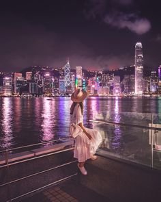 a woman in a dress and hat standing on the edge of a railing overlooking a city at night