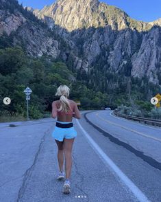 a woman is walking down the road in front of some mountains and trees with her back to the camera