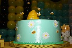 a blue cake decorated with daisies and bees is on a table in front of balloons