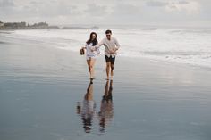 a man and woman walking on the beach with their reflection in the wet sand as they walk towards the water