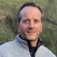 a man wearing a gray sweater and black tie standing in front of some green trees