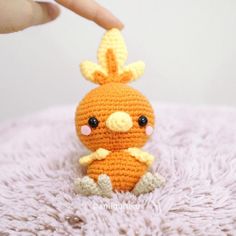 a small crocheted orange bird sitting on top of a white blanket next to a person's hand