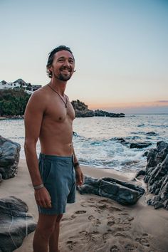 a man standing on top of a sandy beach next to the ocean at sunset with his shirt off