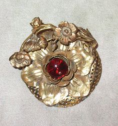 "I think that his lovely antique brooch is from the 20s but it may be from the early 30s.  It measures around 2\" in diameter and the scroll work is raised. The red glass center stone is around 1/2\" in diameter. The BONUS GIFT is a handmade fine wool black bib to wear the pin as a necklace. The bar of the brooch can sit between the layers of the necklace. There is a double fused layer of fabric at the front to support the weight of the pin plus a back lining layer with the name of the designer Antique Red Brooch For Formal Occasions, Vintage Red Brooches For Formal Occasions, Vintage Red Wedding Brooches, Antique Red Brooch As Gift, Antique Red Wedding Brooch, Antique Red Brooches As Gift, Antique Red Brooches As A Gift, Antique Red Brooches For Gift, Vintage Flower Brooch For Evening