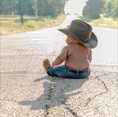 a baby sitting on the ground wearing a cowboy hat and looking at something in the distance