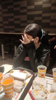 a woman is sitting at a table with food and drinks in front of her, covering her face