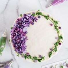 a cake decorated with purple flowers and greenery on a marble countertop next to other decorations