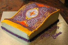 a decorated book cake sitting on top of a silver plate with purple and yellow frosting