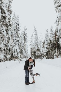 a man and woman kissing in the middle of a snow covered road surrounded by trees