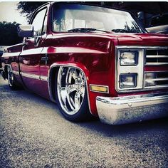 an old red truck parked in a parking lot next to other cars and trucks with chrome rims