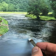 a person is holding a paintbrush in front of a painting of a river and trees