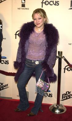 a woman standing on a red carpet with a purple coat over her shoulders and holding a pink purse