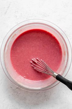 a glass bowl filled with red liquid and a whisk in the middle, on a white surface