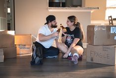 a man and woman sitting on the floor with boxes around them, one holding a beer