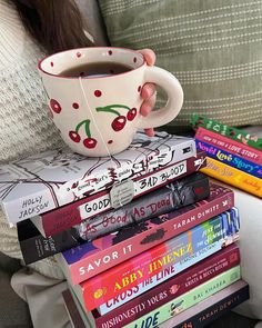 a stack of books and a cup of tea on a couch with a woman's arm holding the mug