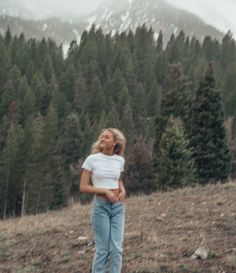 a woman is standing in the middle of a field with trees and mountains behind her