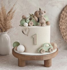 a white cake with teddy bears on top sitting on a wooden stand next to a vase