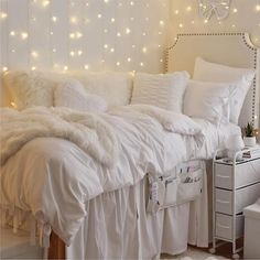 a white bed topped with lots of pillows next to a wall covered in fairy lights