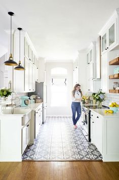 a woman standing in the middle of a kitchen with white cabinets and wood flooring