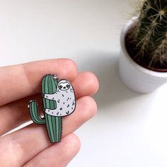 a hand holding a pin with a sloth on it and a cactus in the background