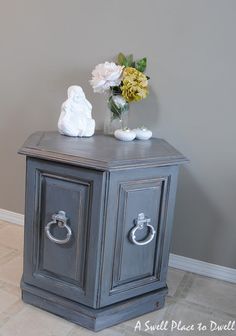 a small gray cabinet with two drawers and flowers on the top, in front of a grey wall