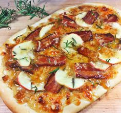 a pizza topped with bacon and apples on top of a wooden cutting board next to a sprig of rosemary