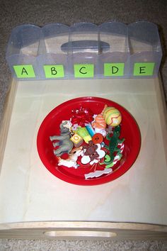 a red bowl filled with plastic toys on top of a wooden table next to an abc letter tray