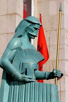 a statue of an egyptian god holding a spear in front of a building with two flags