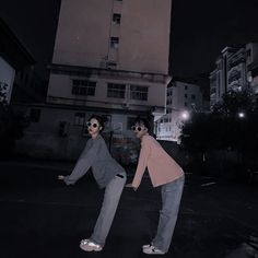 two people standing in the middle of an empty parking lot at night with buildings in the background