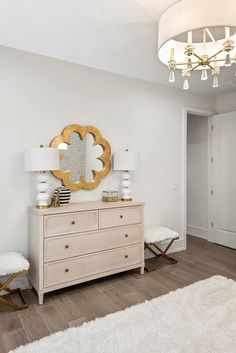 a white and gold bedroom with a dresser, mirror and lights on the sideboard