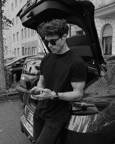 a man standing next to a parked car looking at his cell phone while wearing sunglasses