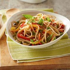 a white bowl filled with noodles and vegetables on top of a wooden cutting board next to a spoon