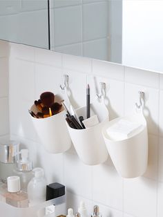 three white cups hanging on the wall above a sink with toothbrushes and other bathroom items