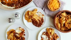 three white plates topped with chili cheese and nachos next to bowls of chips