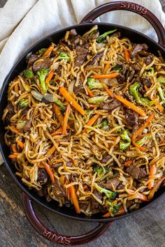 stir fried noodles with beef and vegetables in a skillet on top of a wooden table