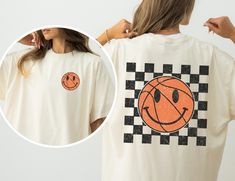 We love Comfort Colors shirts because they are made of 100% ring-spun cotton and are oh so soft. They are garment-dyed and have a vintage feel to them. This one is printed with a retro basketball smiley face on the front and a larger basketball smiley face on the back against checkered board background  If you would like a color you don't see, just reach out! * Fabric weight: 6.1 oz/yd² (206.8 g/m²) * Garment-dyed * Relaxed fit * 7/8″ double-needle topstitched collar * Twill-taped neck and shoul Trendy Crew Neck T-shirt With Smiley Face, Trendy Short Sleeve T-shirt With Smiley Face, Cotton Smiley Face Crew Neck T-shirt, Smiley Face Short Sleeve T-shirt For Streetwear, Fun Cotton T-shirt With Smiley Face, Basketball Mom, Smile Face, Shirt Price, Smiley