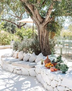 an outdoor seating area under a tree with fruit and vegetables on the stone wall next to it