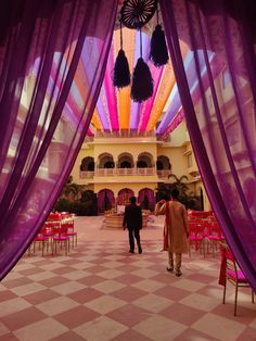 two people walking through an open area with purple drapes on the ceiling and pink chairs