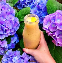 a hand holding a jar filled with liquid next to purple flowers