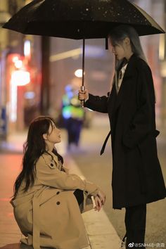 a woman holding an umbrella next to a man sitting on the ground in front of a building