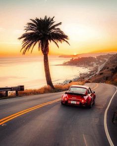 a red sports car driving down the road next to a palm tree and ocean at sunset