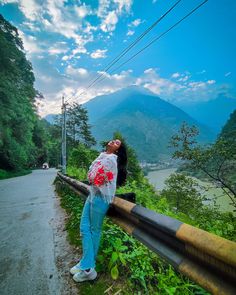 a woman leaning against a rail on the side of a road with mountains in the background