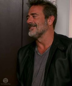 a man with a goatee smiles while standing next to a door and wearing a black leather jacket