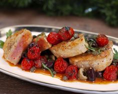 a white plate topped with meat covered in tomatoes and olives on top of a wooden table