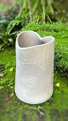 a white vase sitting on top of a green moss covered ground in front of plants